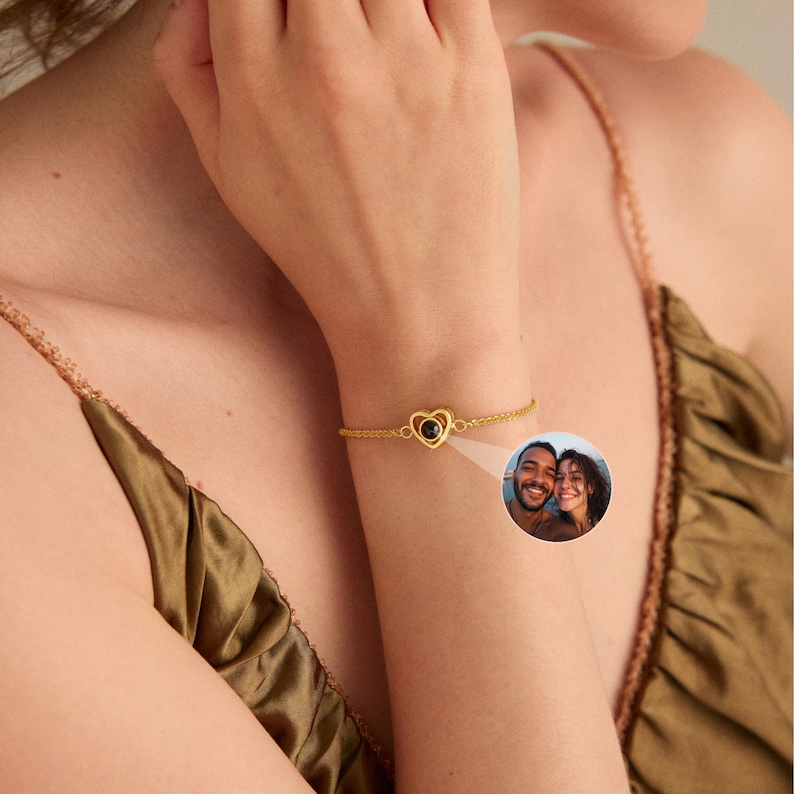May include: A gold-colored bracelet featuring a heart-shaped charm with a black center. The bracelet is worn on a wrist, and a small inset image shows two people. The background is a woman's olive-colored top.