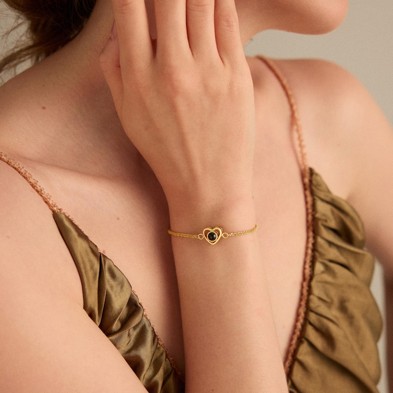 May include: A gold-colored bracelet featuring a heart-shaped pendant with a black stone center. The delicate chain bracelet is worn on a wrist, showcasing the jewelry's design and color against a neutral background.