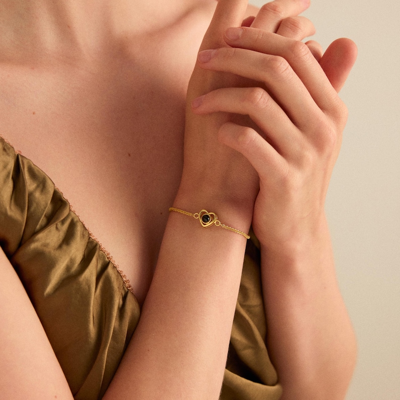 May include: A gold-colored bracelet featuring a heart-shaped pendant with a dark stone. The delicate chain bracelet is worn on a wrist, with the hands of a person in the background. The bracelet is a piece of jewelry.