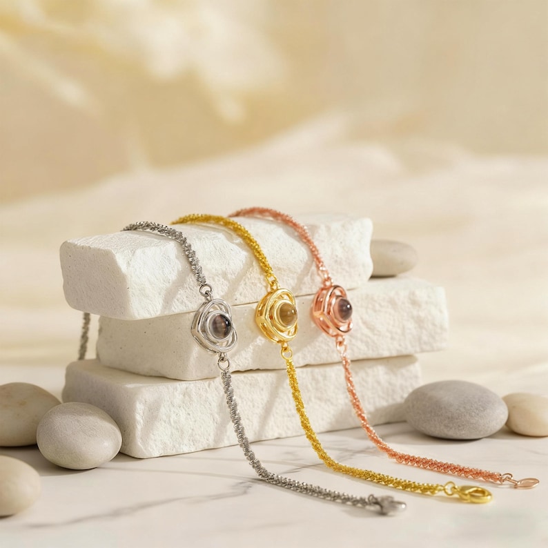 May include: Three delicate bracelets in silver, gold, and rose gold. Each bracelet features a central floral-shaped pendant with a small, round, dark-colored stone. The bracelets are displayed on a stack of white, textured blocks.