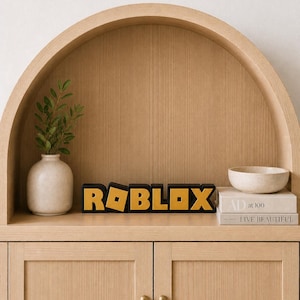 May include: A wooden shelf displays a black and gold "ROBLOX" sign, a small white vase with greenery, a small bowl, and two books. The shelf is part of a light wood cabinet with two doors and brass knobs.