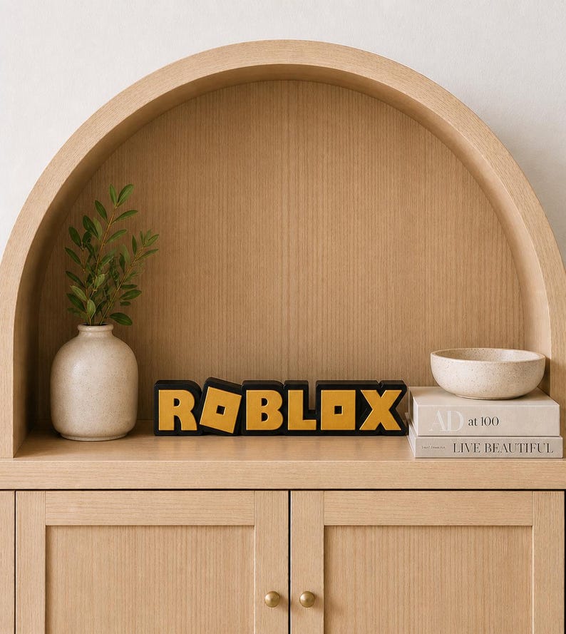 May include: A wooden shelf displays a black and gold "ROBLOX" sign, a small white vase with greenery, a small bowl, and two books. The shelf is part of a light wood cabinet with two doors and brass knobs.