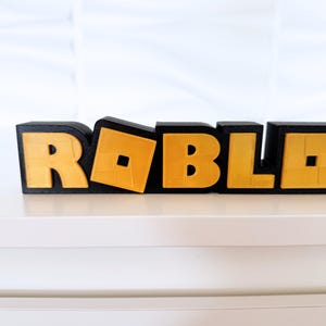 May include: A black and gold 3D-printed sign spells out "ROBLOX". The letters are black with gold-colored infill. The "O" is tilted, adding a dynamic element to the design. The sign is displayed on a white surface.