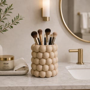 May include: A beige, bubble-textured makeup brush holder filled with various brushes. The holder sits on a marble countertop next to a jar of OUAI hair treatment and a small white towel. A gold-framed mirror and faucet are in the background.