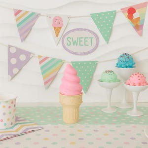 Può includere: Una scena festosa con coni gelato e ghirlande decorative. Un cono gelato rosa a spirale è in primo piano. Striscioni dai colori pastello e decorazioni a forma di gelato sono appesi sopra. La parola "SWEET" è visualizzata su un cartello ovale.