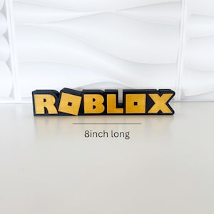 May include: A black and gold Roblox logo, 8 inches long. The word "ROBLOX" is displayed in a bold, blocky font with gold lettering and a black outline. The background is a white, textured surface.
