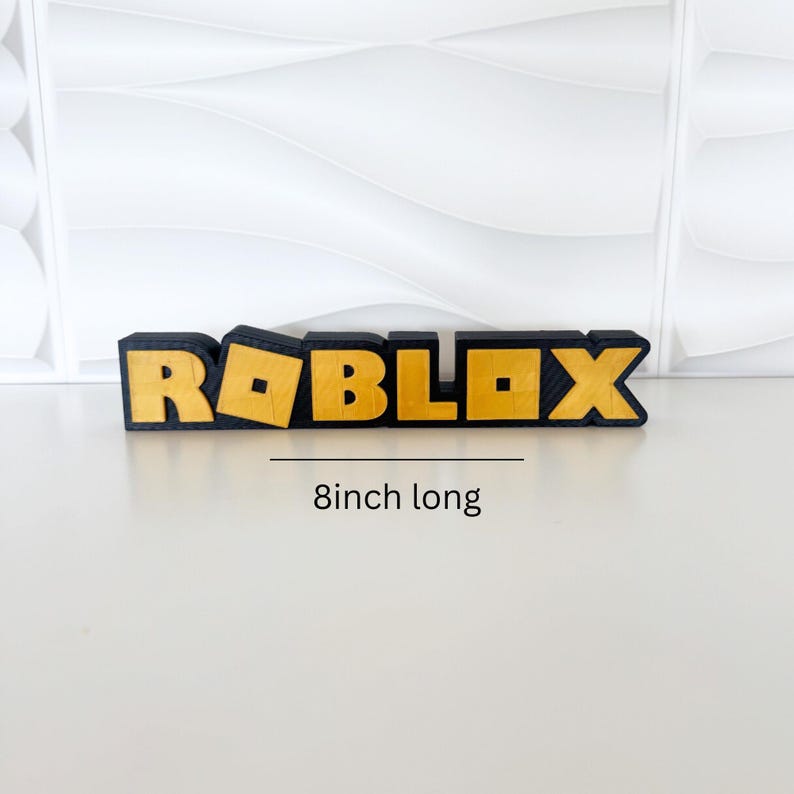 May include: A black and gold Roblox logo, 8 inches long. The word "ROBLOX" is displayed in a bold, blocky font with gold lettering and a black outline. The background is a white, textured surface.
