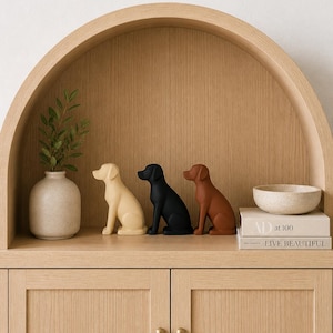 Peut inclure: Trois figurines de chiens, crème, noir et marron, sont disposées sur une étagère en bois. Un vase avec de la verdure, un petit bol et deux livres sont également présents. L'étagère fait partie d'un meuble en bois.