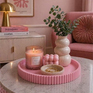 Puede incluir: Una bandeja decorativa rosa contiene una vela encendida, una pequeña vela rosa con forma de burbuja, un cuenco pequeño con una pulsera dorada y un jarrón beige con vegetación. La vela está en un recipiente de vidrio transparente con el texto "VOLUSPA ROSE OTTO".