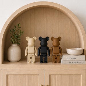 Könnte beinhalten: Drei Bearbrick-Figuren in Beige, Schwarz und Braun stehen auf einem Holzregal. Die Figuren haben ein blockartiges Design mit bärenartigen Ohren. Eine kleine Vase mit Grünzeug und eine Schale befinden sich ebenfalls auf dem Regal.