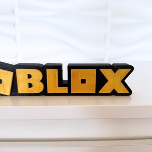 May include: A 3D-printed sign spelling out "ROBLOX" in black and gold. The letters are blocky and textured, with the gold sections appearing to have a metallic finish. The sign is placed on a white surface.