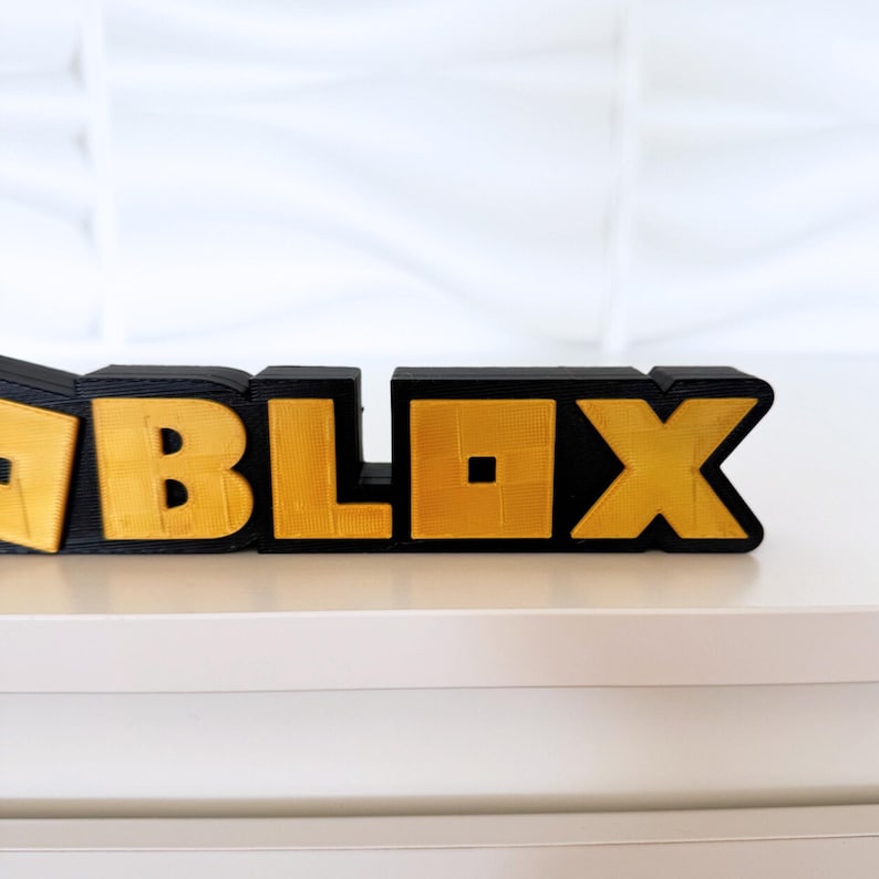 May include: A 3D-printed sign spelling out "ROBLOX" in black and gold. The letters are blocky and textured, with the gold sections appearing to have a metallic finish. The sign is placed on a white surface.