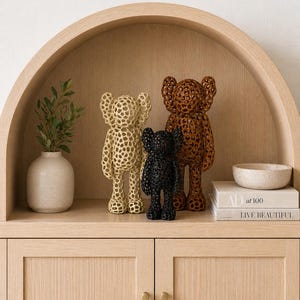 May include: Three bear-shaped decorative figurines in cream, black, and brown, featuring a perforated design. They are displayed on a wooden shelf with a vase of greenery, a small bowl, and two books.