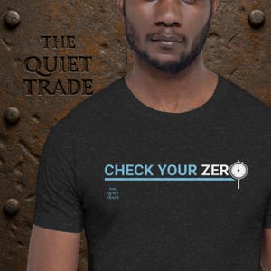 May include: Dark gray t-shirt with the text "CHECK YOUR ZERO" in light blue and a small clock graphic. The shirt also features the text "THE QUIET TRADE" in white.