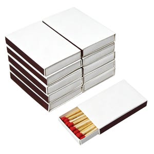 May include: A stack of five rectangular matchboxes with plain white sliding covers and dark brown inner trays. One additional matchbox is positioned horizontally in front, partially open to reveal wooden matches with bright red tips.