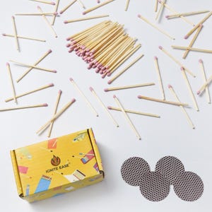 May include: A collection of wooden matches with pink tips scattered on a white surface. A yellow box with the text "IGNITE EASE" and three circular match strikers are also present. The matches are for lighting candles or other items.