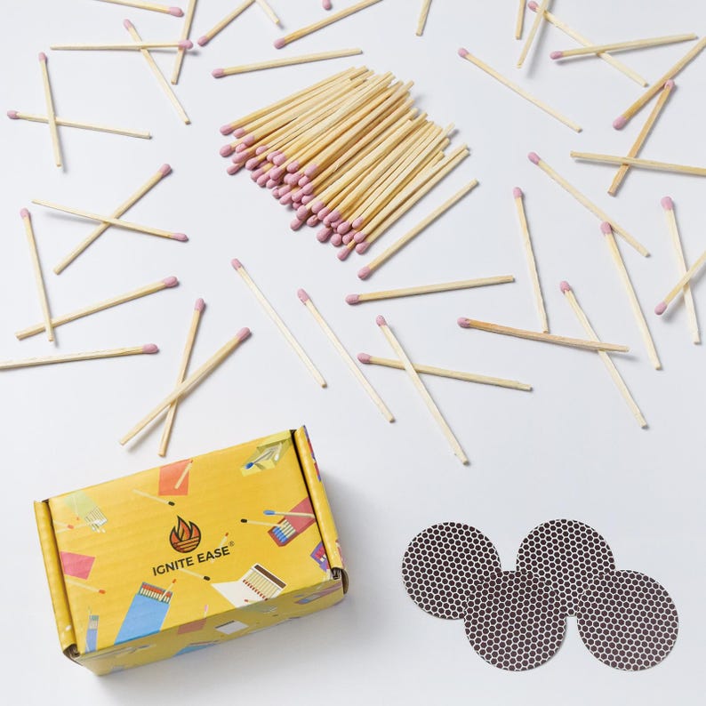 May include: A collection of wooden matches with pink tips scattered on a white surface. A yellow box with the text "IGNITE EASE" and three circular match strikers are also present. The matches are for lighting candles or other items.