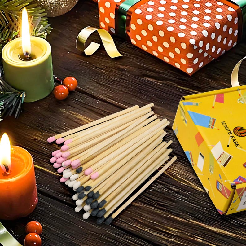 May include: A collection of wooden matches with pink, white, and black tips, arranged on a dark wooden surface. Two lit candles and a gift box with a gold ribbon are in the background. A yellow box with the text "IGNITE EASE" is also visible.