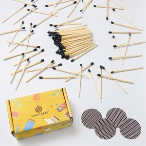 May include: A collection of wooden matches with black tips scattered on a white surface. A yellow box with the text "IGNITE EASE" and a flame logo is visible, along with three brown match striker pads. The matches are a light brown color.