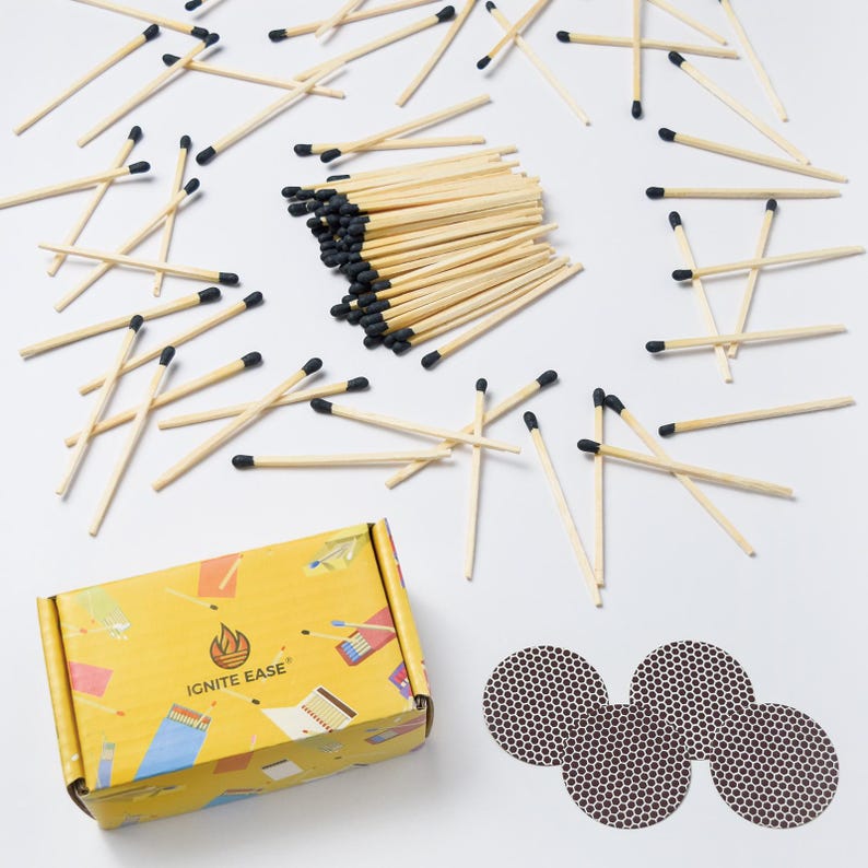 May include: A collection of wooden matches with black tips scattered on a white surface. A yellow box with the text "IGNITE EASE" and a flame logo is visible, along with three brown match striker pads. The matches are a light brown color.