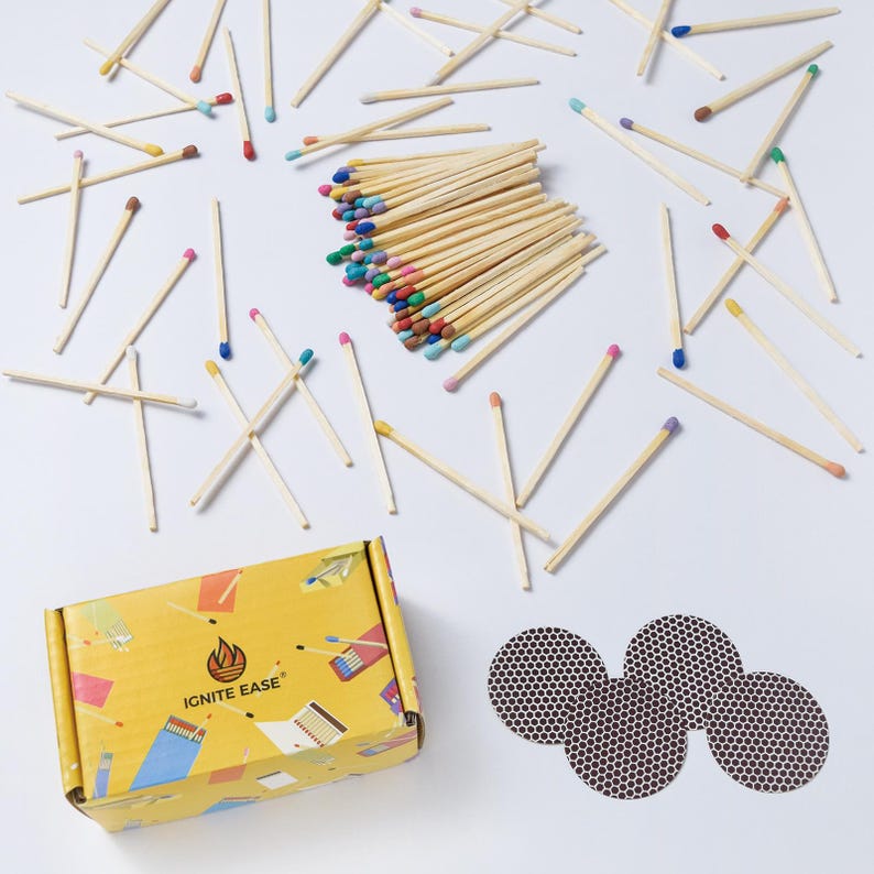 May include: A collection of wooden matches with colorful tips scattered on a white surface. A yellow box with the text "IGNITE EASE" and a flame logo is visible, along with three round match strike pads.