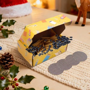 May include: A yellow matchbox with a colorful design, open to reveal numerous wooden matches with blue tips. A strip of match striker pads is next to the box. The scene is set on a cream-colored knitted surface, with holiday decorations in the background.