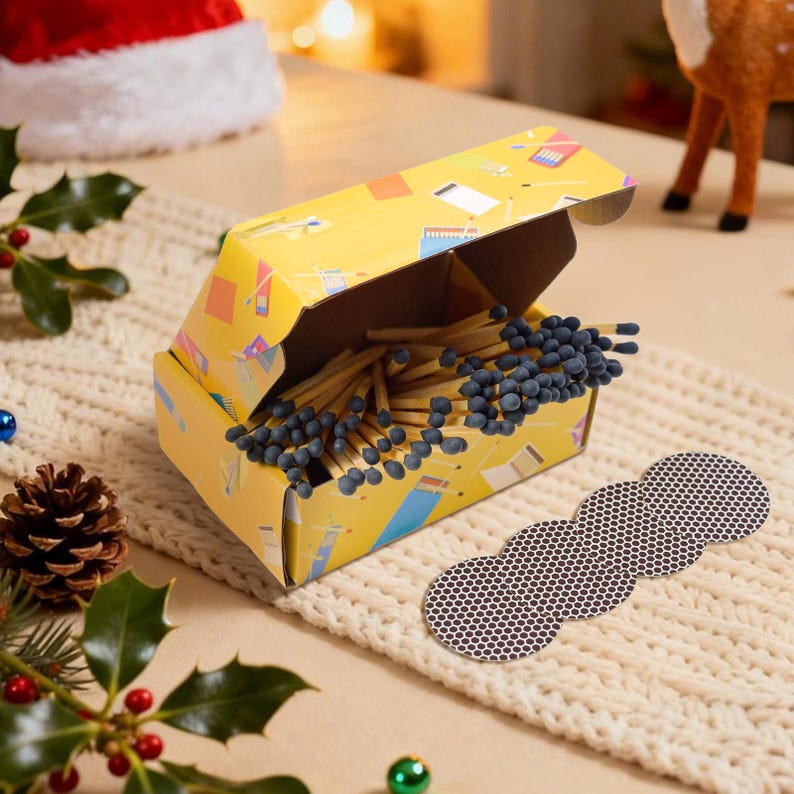 May include: A yellow matchbox with a colorful design, open to reveal numerous wooden matches with blue tips. A strip of match striker pads is next to the box. The scene is set on a cream-colored knitted surface, with holiday decorations in the background.