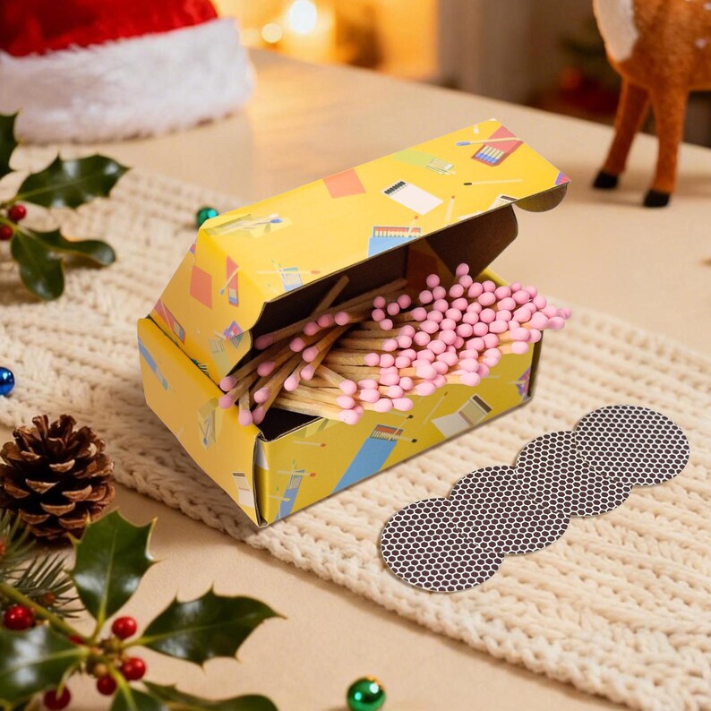 May include: A yellow matchbox with a colorful design, filled with pink-tipped matches. A brown, scalloped match striker is next to the box. The scene includes holiday decorations, such as holly and a Santa hat, suggesting a festive theme.