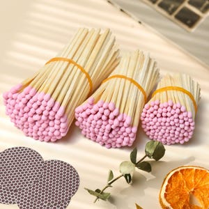 May include: Three bundles of wooden matches with light pink heads, each secured by an orange rubber band. Included are three circular dark brown striker pads with a honeycomb pattern and a dried orange slice with a green eucalyptus sprig.