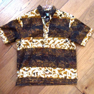 May include: A short-sleeved shirt featuring a dark brown, tan, and cream-colored batik-style pattern. The garment has a pointed collar, a partial button-down placket at the center front, and a repeating horizontal design of abstract shapes.