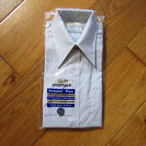 May include: A new, folded button-down shirt in a clear plastic package. The shirt is light gray with a repeating pattern and a pointed collar. The packaging includes the text "WESTVALE Permanent Press" and "NEVER NEEDS IRONING".