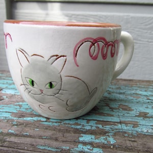 May include: White ceramic mug with a cat illustration. The mug features a gray cat with green eyes and pink swirls. The rim is brown, and the handle is white. A charming and whimsical design for cat lovers.