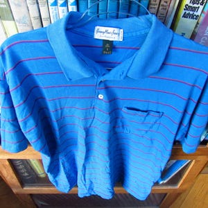 May include: Blue polo shirt with red horizontal stripes. The shirt has a pocket on the left chest and a three-button placket. The collar is a darker shade of blue. The shirt is hanging on a hanger.