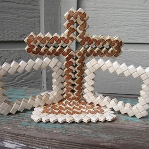 May include: A decorative sculpture constructed from folded paper or plastic strips, featuring a central cross shape flanked by two rectangular loops. The object displays a repeating pattern of alternating white and brown textured segments throughout.