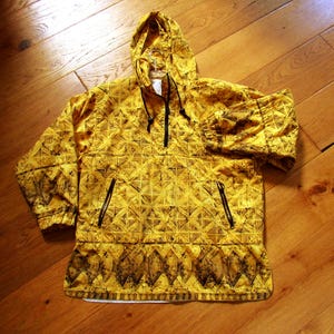 May include: A yellow hooded anorak with a geometric pattern in black. The anorak has a quarter-zip closure, two front pockets with black zippers, and a drawstring hood. The garment is made of a lightweight material.