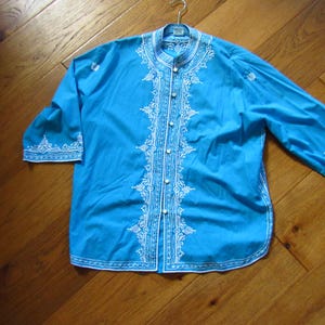 May include: A turquoise shirt with white embroidered detailing. The shirt features a mandarin collar, button-down front, and three-quarter sleeves. White embroidery adorns the collar, front, and sleeve cuffs. The shirt is hanging on a metal hanger.