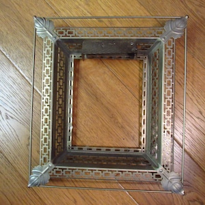 May include: A vintage metal picture frame with a rectangular shape. The frame has a decorative, openwork design with leaf accents at the corners. The frame is made of a light-colored metal and is set against a wooden surface.