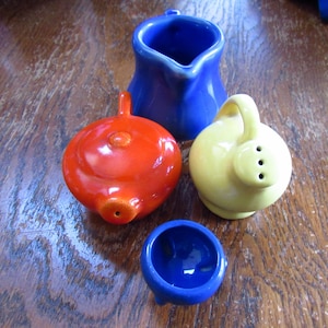 May include: A collection of colorful ceramic kitchenware, including a cobalt blue creamer, a bright orange condiment holder, a yellow salt shaker, and a small blue bowl. The pieces are arranged on a wooden surface.