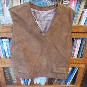 May include: A brown suede vest featuring a V-neckline, a smooth satin-like lining in a matching light brown tone, and two rectangular flap pockets positioned at the lower front section of the garment.