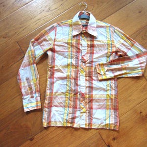 May include: A long-sleeve button-down shirt featuring a plaid pattern in shades of orange, yellow, white, and light blue. The shirt has a pointed collar, a front button placket, and buttoned cuffs on the sleeves, presented on a gray hanger.