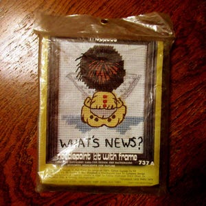 May include: A sealed needlepoint kit with a frame. The kit features a cartoon character with a spiky brown hair and a yellow outfit. The text "WHAT'S NEWS?" is visible. The kit is packaged in a clear plastic bag.