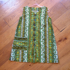 May include: A sleeveless dress with a vibrant, patterned design in shades of green, white, and gold. The dress features a button-down front, two front pockets, and a unique vertical stripe pattern. The dress is hanging on a pink hanger.