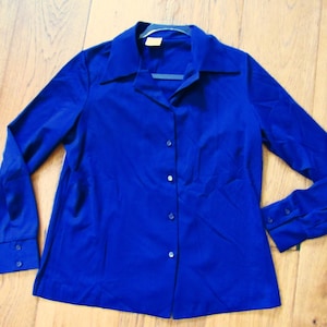 May include: A long-sleeved, cobalt blue button-down shirt with a classic collar. The shirt has buttoned cuffs and a tailored fit. The fabric appears to be a smooth, possibly synthetic material. The shirt is displayed on a wooden surface.