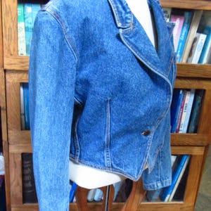 May include: A blue denim jacket with a classic design. The jacket features a notched collar, long sleeves, and a single-button closure. The jacket is displayed on a mannequin and has visible stitching details.