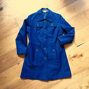 May include: A solid royal blue coat featuring a classic collar, long sleeves, and a front button closure. The garment includes two rectangular chest pockets with button flaps and two vertical hip pockets, all accented with matching blue buttons.