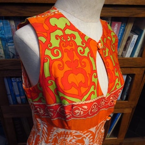 May include: A sleeveless dress featuring a vibrant orange and lime green damask-style pattern on the bodice. The garment has a deep keyhole neckline secured by a single orange button at the top and a horizontal band with a floral border design.