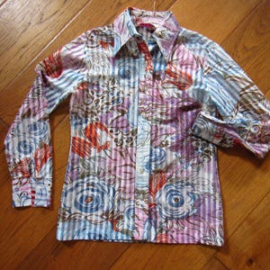 May include: A button-down shirt with a colorful floral pattern. The shirt features long sleeves and a collared neckline. The design includes stripes, flowers, and abstract shapes in shades of blue, red, purple, and white.