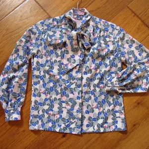May include: A long-sleeved blouse with a floral pattern in blue, gray, and pink. The blouse has a bow tie collar and buttoned cuffs. The fabric appears to be lightweight, and the blouse is hanging on a hanger.