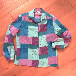 May include: A long-sleeved button-down shirt featuring a patchwork design of rectangular fabric panels in shades of teal, light blue, and magenta. Each panel displays a small, repeating floral pattern. The shirt has a collar and white buttons.