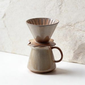 May include: A ceramic pour-over coffee maker in shades of brown and beige. The set includes a cone-shaped dripper with vertical ridges, a flower-shaped base, and a small jug with a handle. The coffee maker is designed for manual brewing.